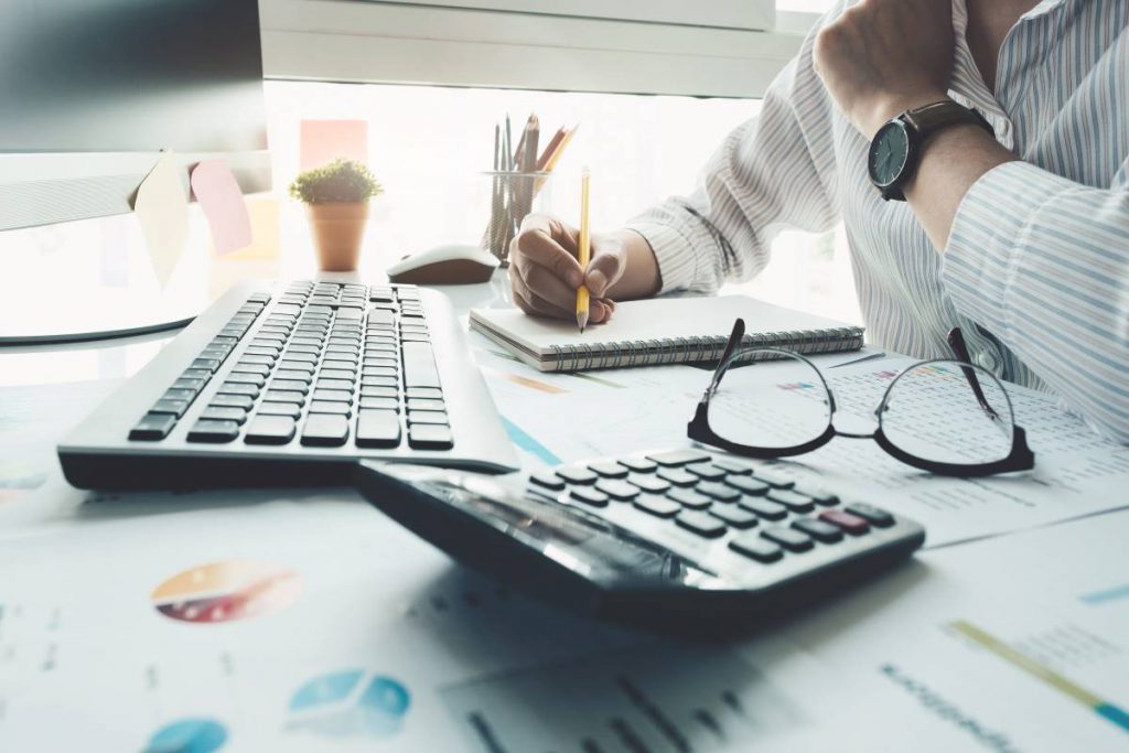 Uma pessoa sentada à mesa de trabalho com um teclado, óculos, uma calculadora e documentos financeiros, escrevendo notas, simbolizando tarefas relacionadas à gestão financeira como orçamento, bancos e impostos.