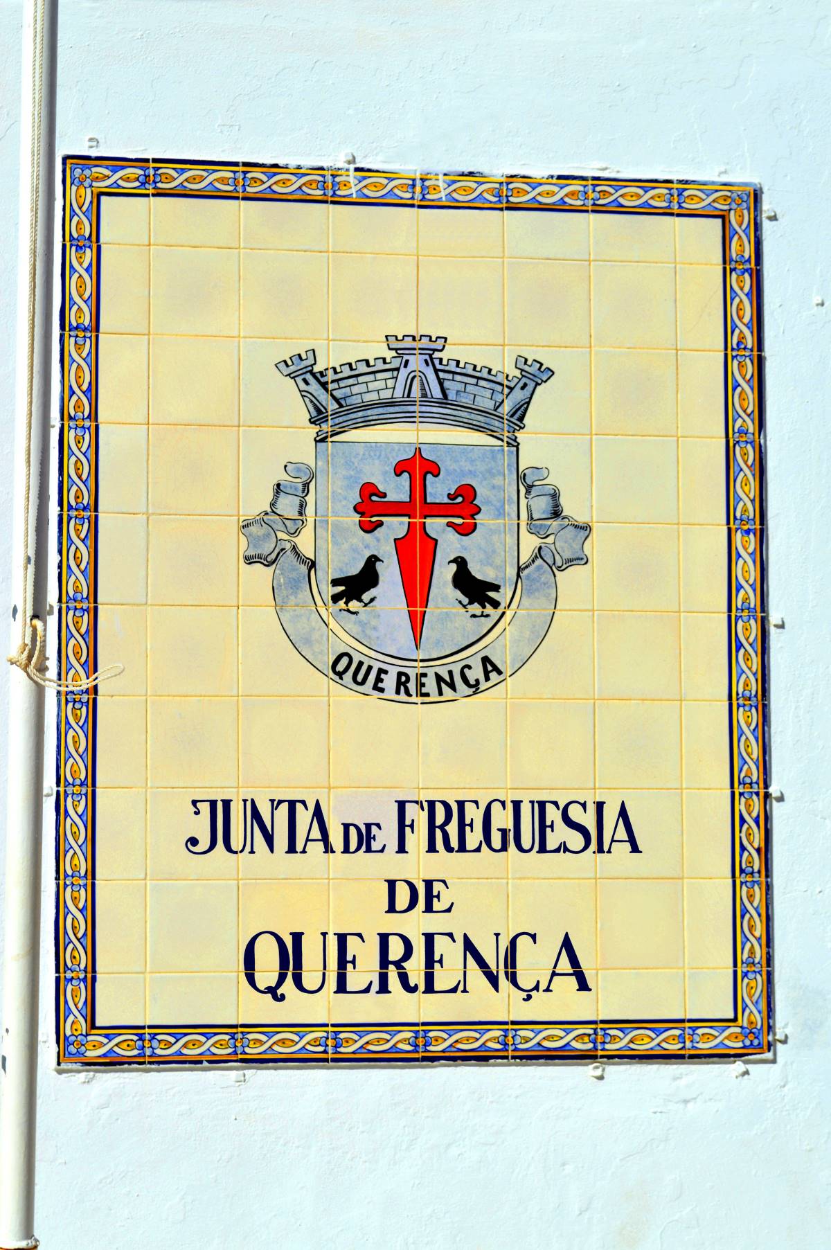Placa de azulejos tradicional portuguesa na parede indicando "Junta de Freguesia de Querença", simbolizando a administração local e serviços ao cidadão, incluindo a confirmação de morada e orientações para mudança de residência.
