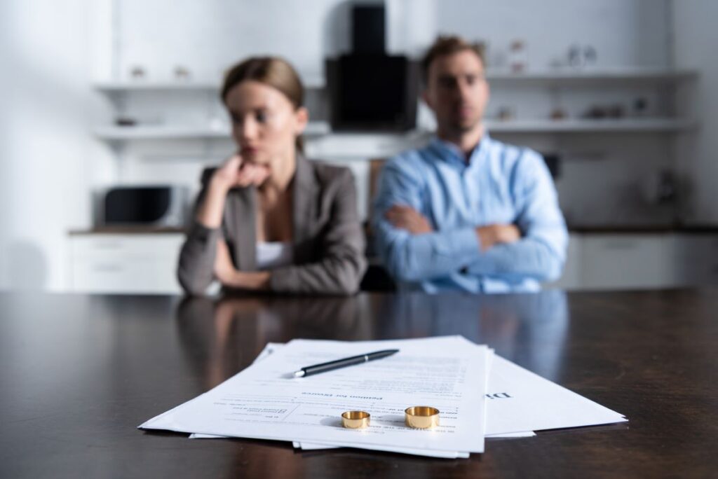Um casal sentado em lados opostos de uma mesa, com uma certidão de divórcio, uma caneta e alianças de casamento colocadas em destaque no primeiro plano.
