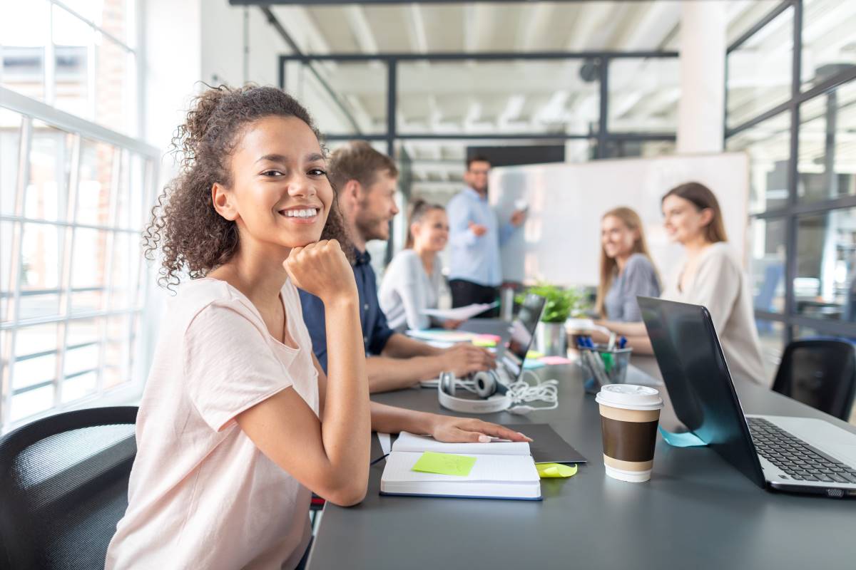 Uma jovem profissional sorridente senta-se à mesa de uma sala de reuniões moderna e iluminada, rodeada de colegas e um laptop aberto. Ela exibe uma expressão de confiança e satisfação, representativa da juventude qualificada em Portugal que é incentivada a permanecer no país graças ao novo prémio de incentivo lançado em 2024.