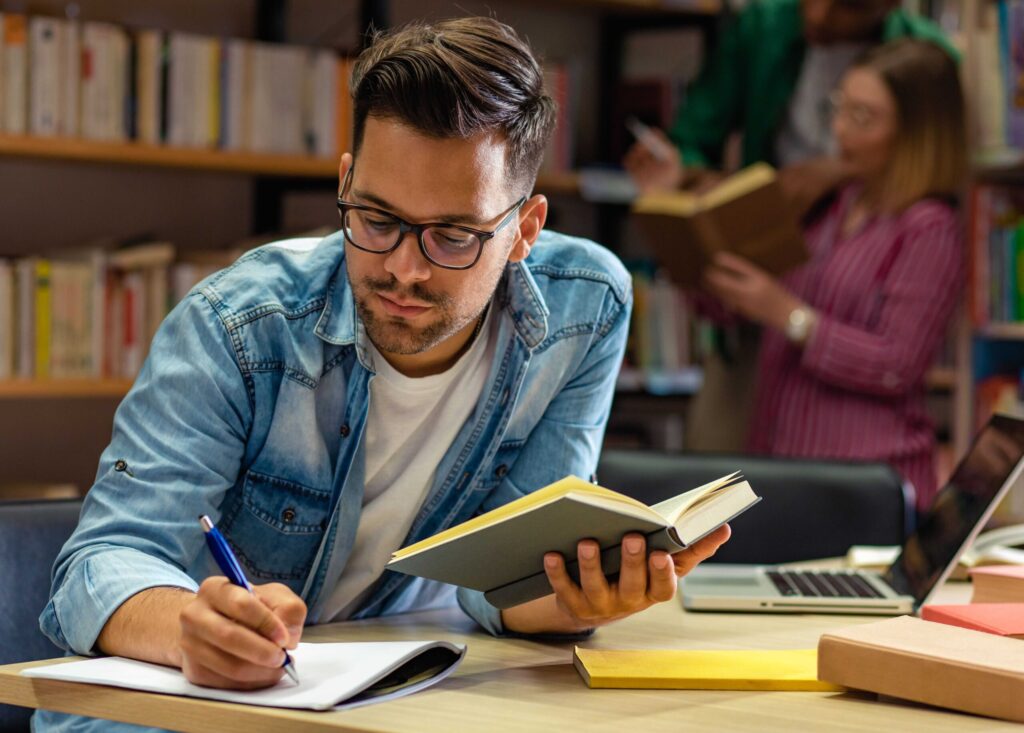 Jovem estudante sentado em uma biblioteca, lendo um livro e fazendo anotações.